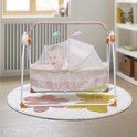 Een baby ligt in een schommelwieg met klamboe. De wieg is versierd met een bloemenrand en hangende speelgoedjes.