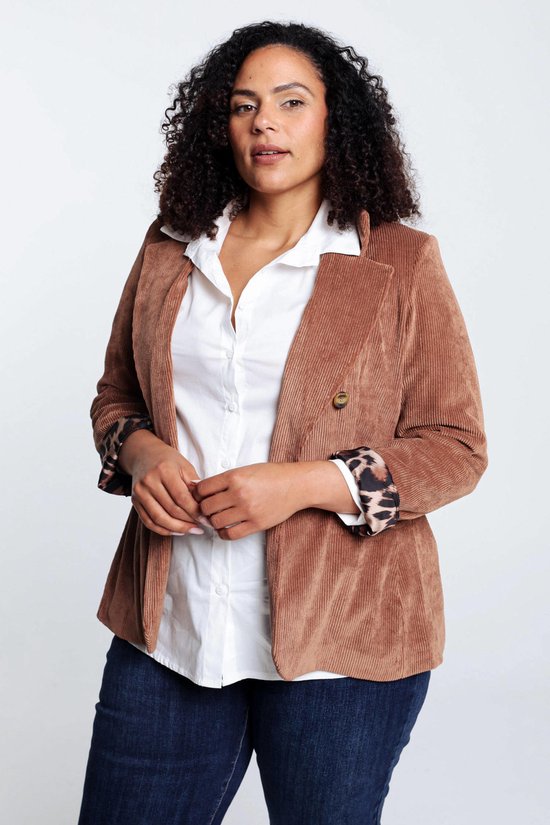 Vrouw met krullend haar in een bruine corduroy blazer met luipaardprint manchet, witte blouse en donkere jeans.