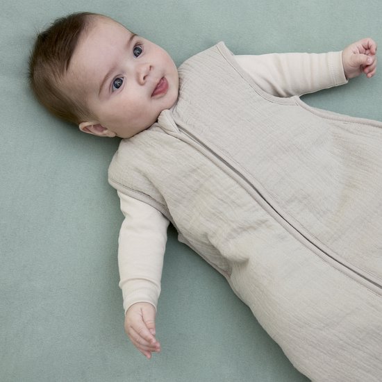 Baby ligt op de rug in een turquoise bedje, draagt een beige slaapzak.