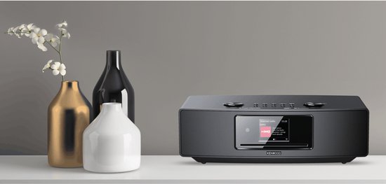 Zwarte Kenwood radio op een witte plank, naast gouden en zwarte decoratieve vazen met witte bloemen.