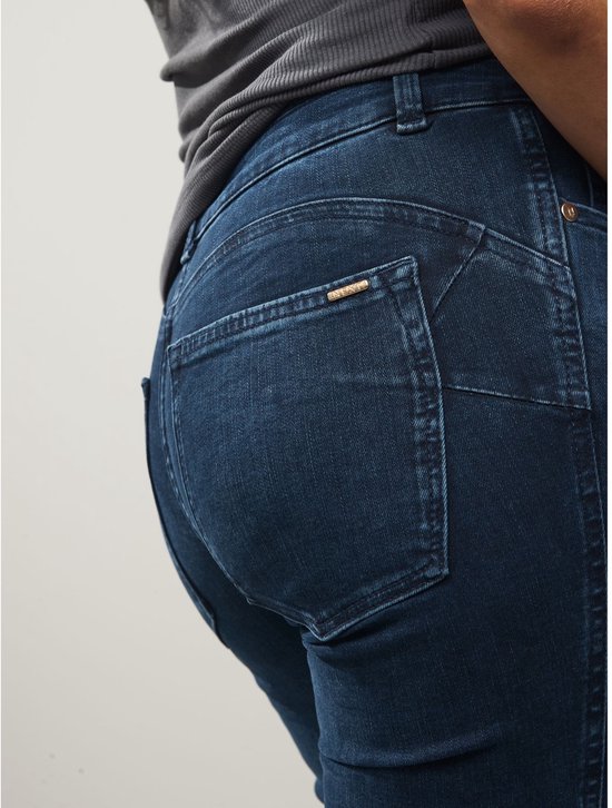 Close-up van de achterkant van een donkerblauwe spijkerbroek met bootcut pasvorm. De broek heeft opgestikte achterzakken met een metalen detail.