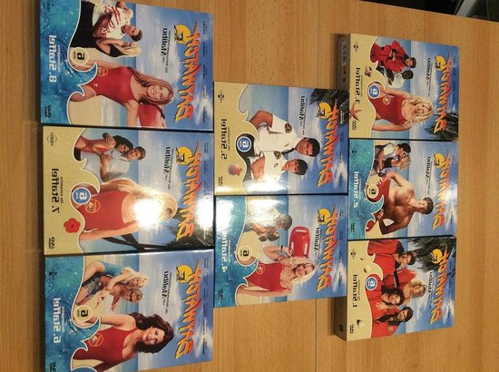 Meerdere dvd-boxen van de serie Baywatch met beelden van reddingsacties op het strand, mannen en vrouwen in rode badpakken.
