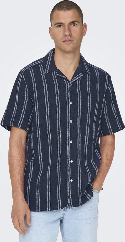 Man van voren, draagt een donkerblauw gestreept kortsvormig overhemd en een lichtblauwe jeans.