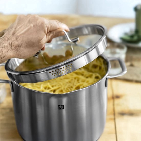Een hand tilt het glazen deksel met geperforeerde rand van een roestvrijstalen kookpot gevuld met spaghetti.