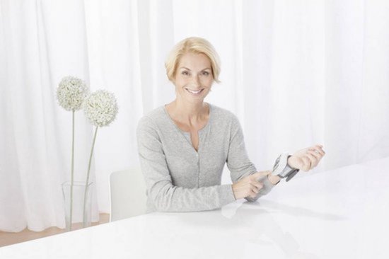 Vrolijke blonde vrouw met grijze vest, die haar polsbloeddrukmeter laat zien.