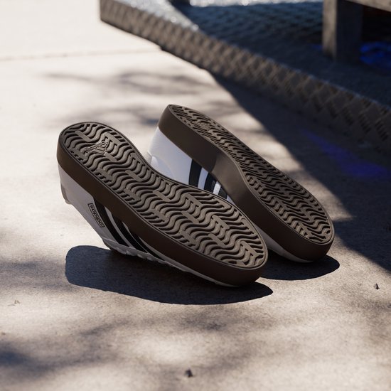Witte Adidas sneakers met zwarte strepen, bruine zolen, op de grond.