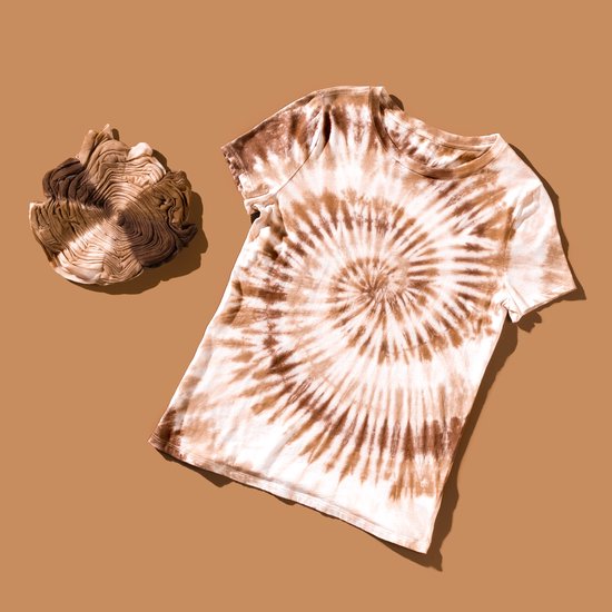Jonge man met zonnebril, leunt tegen een gele bakstenen muur, draagt een bruin-witte tie-dye t-shirt.