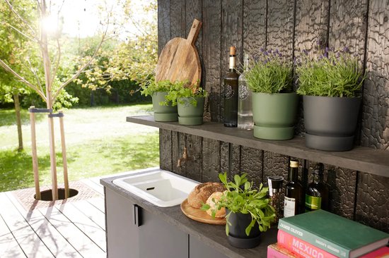 Olijfgroene en terracotta potten met planten op een houten plank, naast een aanrecht.