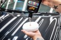 Hand met tatoeages met roze polijstmiddel erop, op een witte applicator, die wordt aangebracht op een glanzende zwarte auto.
