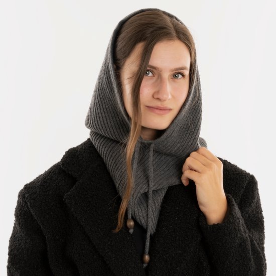 Een vrouw met lang bruin haar, draagt een donkere jas en een grijze gebreide colsjaal die als capuchon is gedragen.