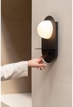 Hand drukt op de schakelaar van een zwarte wandlamp met ovale melkglazen kap.