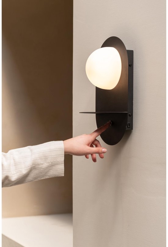 Hand drukt op de schakelaar van een zwarte wandlamp met ovale melkglazen kap.
