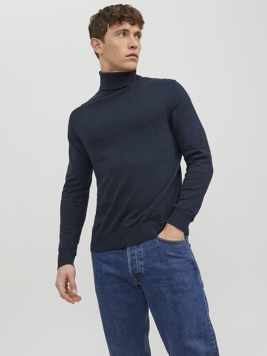 Man met een donkerblauwe coltrui en spijkerbroek, hand in de zak.
