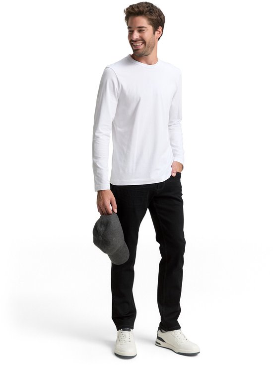Man in zwarte jeans en witte longsleeve, met pet in hand.