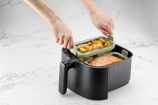 Lichtgroene glazen airfryer schalen, één met zalm en broccoli, de ander met aardappelpartjes, geplaatst in een zwarte airfryer.