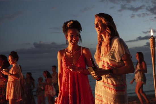 Twee lachende vrouwen op het strand bij zonsondergang; één in rode jurk, de ander in gestreept jurkje met fles.