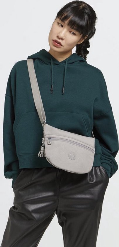 Model met een donkergroene hoodie en zwarte broek draagt een lichtgrijze Kipling schoudertas.
