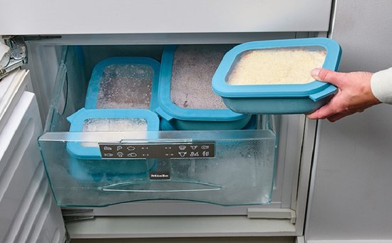 Main retire une boîte de conservation Mepal bleue avec du riz d'un congélateur au contenu glacé.