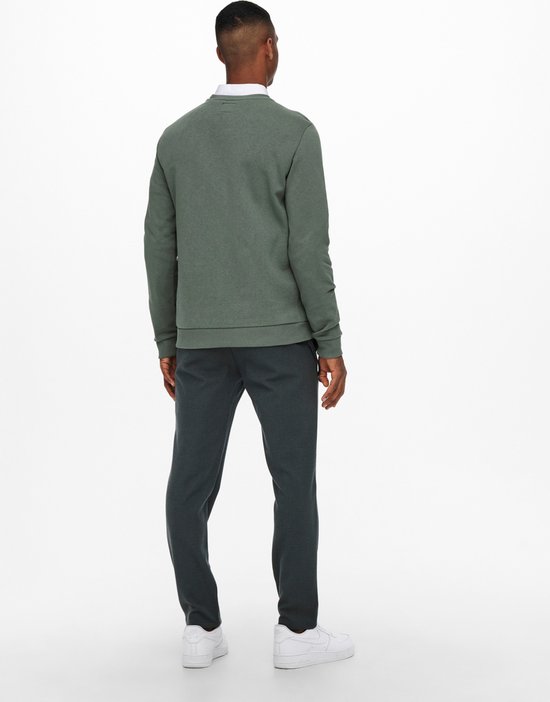 De achterkant van een man met een witte overhemdkraag en mosgroene sweater, gekleed in een grijze broek met ruitpatroon.