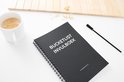 Zwart notitieboek, kopje koffie, papieren clips, bril en pen op een witte tafel.