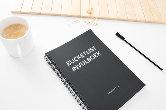 Zwart notitieboek, kopje koffie, papieren clips, bril en pen op een witte tafel.