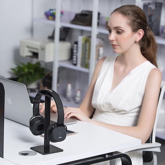 Zwarte koptelefoonstandaard met een zwarte koptelefoon erop, geplaatst op een bureau met een laptop en een vrouw op de achtergrond.