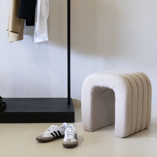 Een beige, bouclé poef met verticale ribbels staat naast een kledingrek met jas en witte blouse en een paar sneakers.