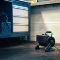 Zwarte keramische kachel met turkooizen handvat en steunen, staand op een werkplaatsvloer naast een gereedschapskast.