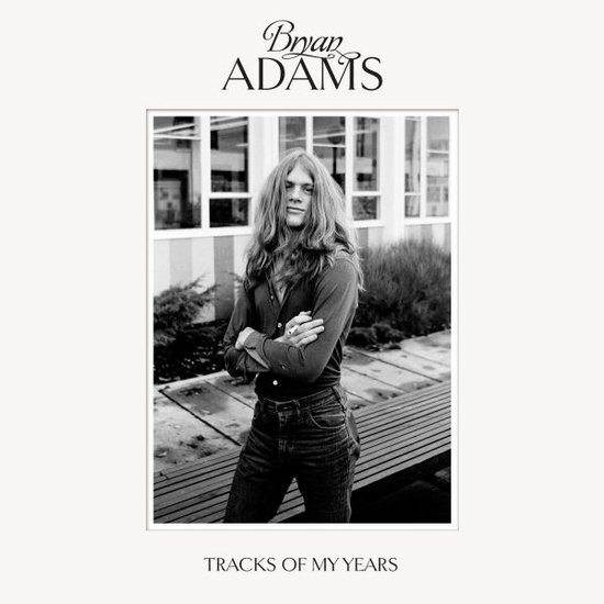 Bryan Adams staat met gekruiste armen voor een gebouw met grote ramen. 'Bryan Adams Tracks of My Years'.