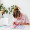 Fille avec un chignon portant un t-shirt rose, collant des autocollants dans un livre d'activités avec des licornes et un arc-en-ciel. 'Holiday' sur le t-shirt.