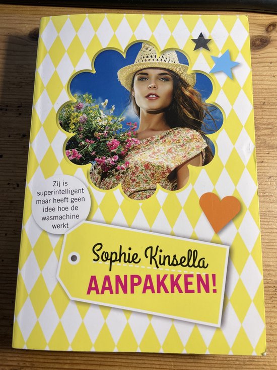 Boekomslag met gele ruitjes. Een jonge vrouw met strohoed, bloemig topje, kijkt lachend. De titel 'Sophie Kinsella AANPAKKEN!' staat op een gele label.