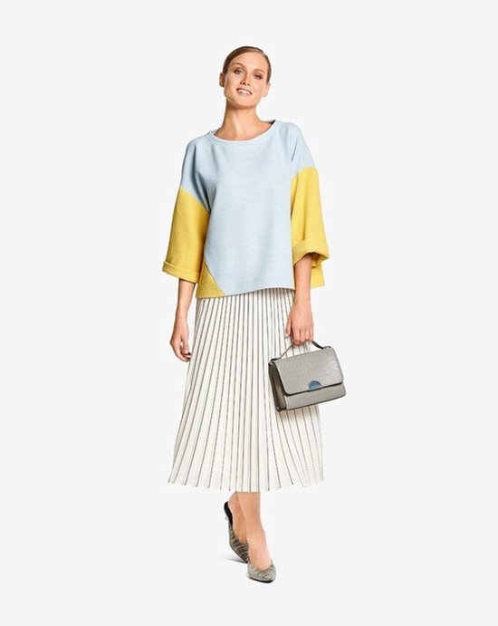 Model draagt een lichtblauwe en gele trui met wijde mouwen en een witte geplooide rok met verticale strepen.