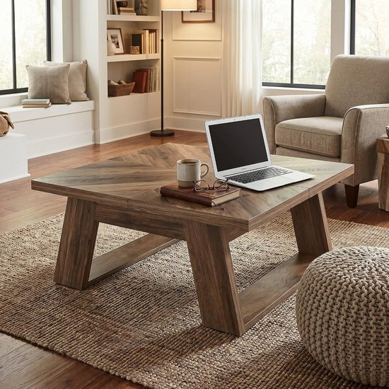 Vierkante salontafel met houten blad in visgraatpatroon en U-vormige poten, met een laptop, mok en bril erop, in een woonkamer met fauteuil.