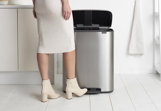Vrouw in beige jurk en laarzen opent een mat stalen Brabantia prullenbak met twee compartimenten.
