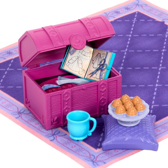 Roze kist met accessoires voor poppen: een modeboek, schaar, stof, kussens, kopje en gebakjes.