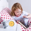 Een meisje leest een boek in bed, naast een witte schaapwekker met gele wijzerplaat. De wekker toont 6:45.