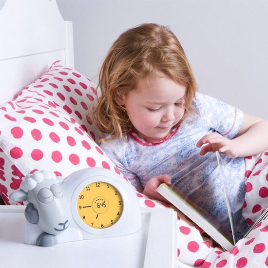 Een meisje leest een boek in bed, naast een witte schaapwekker met gele wijzerplaat. De wekker toont 6:45.