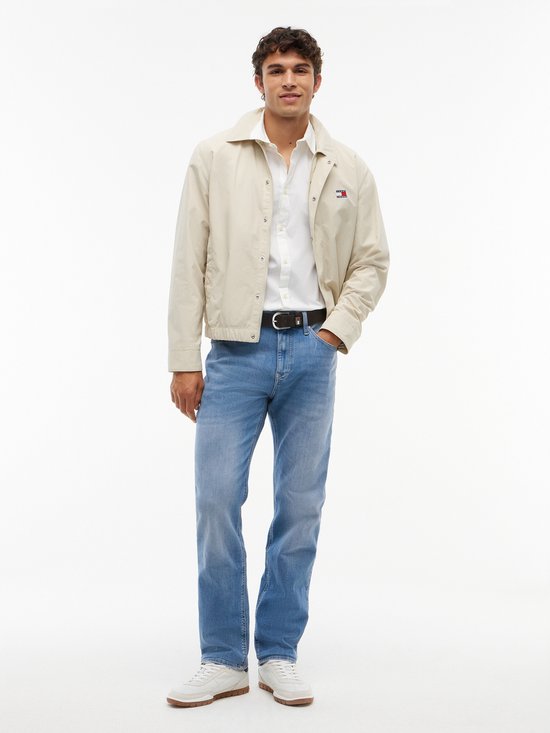 Man in een lichtbeige Tommy Jeans jas, wit overhemd, spijkerbroek en witte sneakers. Logo op de jas.