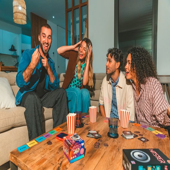 Vier lachende vrienden spelen Hitster rond een houten tafel met popcorn en kaarten. De speldoos is zichtbaar.