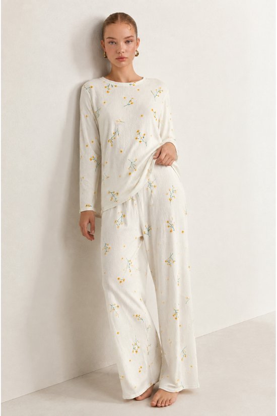 Model draagt een ecru pyjamaset met gele bloemenprint, bestaande uit een shirt met lange mouwen en een wijde broek.