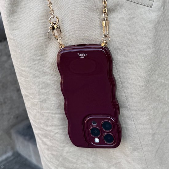 Bordeauxrood glanzend golvend iPhone-hoesje met gouden ketting, gedragen over een beige broek.