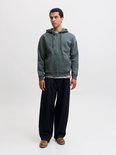 Model in een effen, donkergroene hoodie met rits, zwarte broek en bruine schoenen.