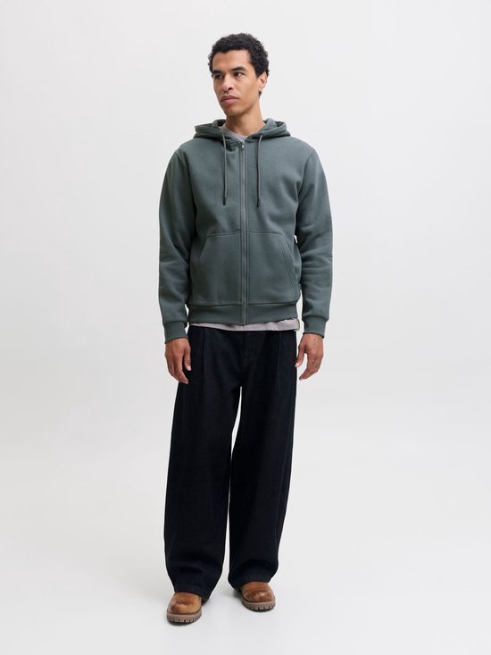 Model in een effen, donkergroene hoodie met rits, zwarte broek en bruine schoenen.