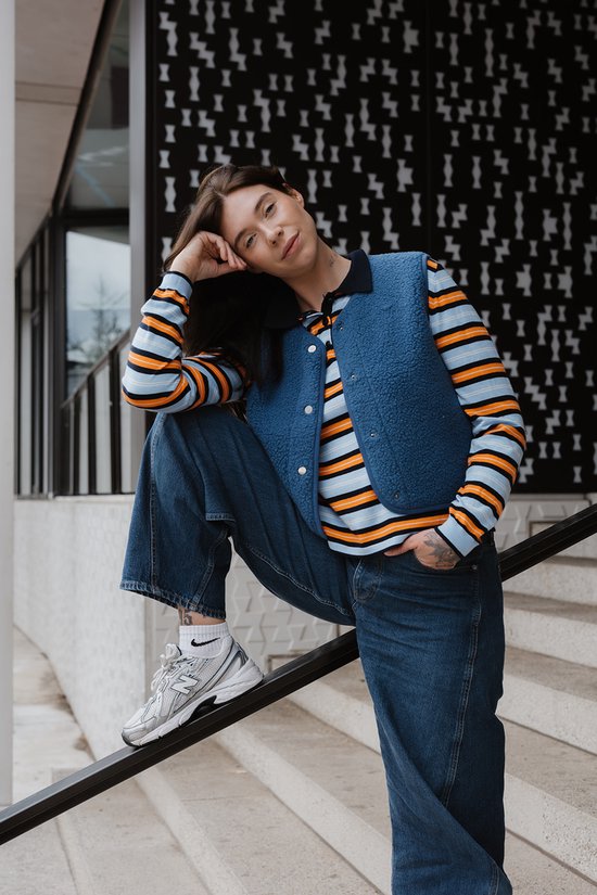 Model zittend, draagt een donkerblauwe teddy bodywarmer over een gestreept shirt, met jeans.