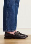 Zwarte leren loafers met een goudkleurig bitdetail gedragen onder een blauwe spijkerbroek.