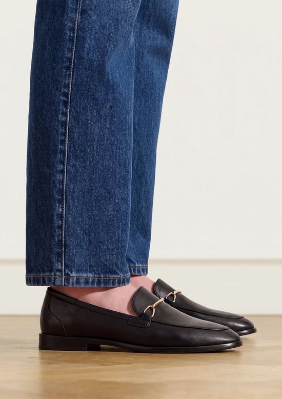 Zwarte leren loafers met een goudkleurig bitdetail gedragen onder een blauwe spijkerbroek.