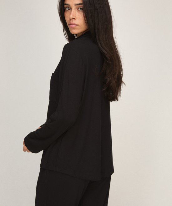 Model met donker haar draagt een zwart pyjamajasje met kraag en borstzak, zijaanzicht.