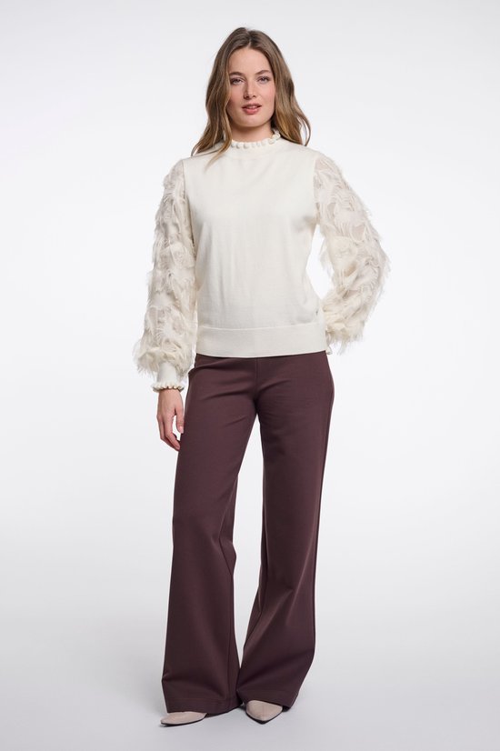 Staand model in een beige trui met franje-mouwen en een donkerbruine pantalon met wijde pijpen.