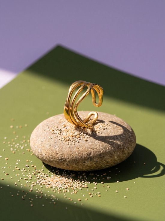 Goudkleurige, gelaagde open ring met golvend design op een steen met zand, tegen een groen en paars achtergrond.