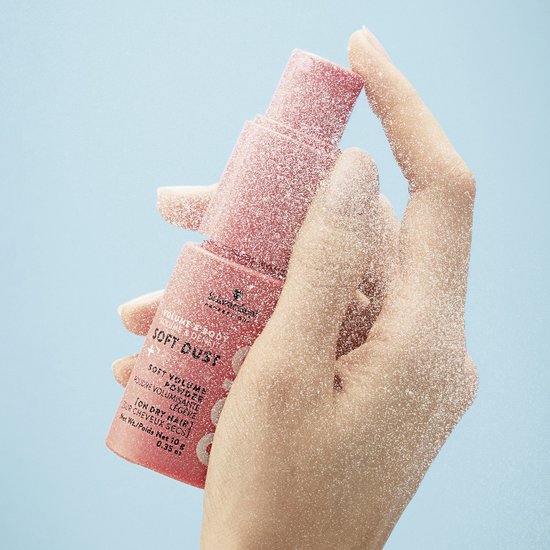 Roze Schwarzkopf OSiS+ Soft Dust volumepoederfles vastgehouden door een hand, omringd door poederdeeltjes.